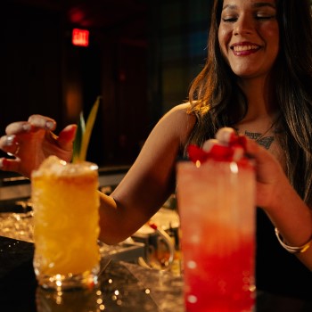 Houndsmen Lounge female bartenders garnishing a margarita and strawberry daiquiri in tall glasses.