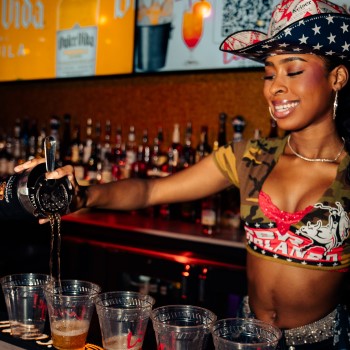 Female bartender behind the bar of PBR Orlando wearing patriotic cowboy hat and cropped top pouring drinks from a cocktail shaker into a line of clear plastic, Live! branded cups.