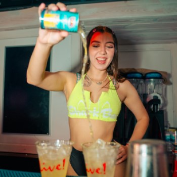 Female bartender in neon yellow tank pouring canned pineapple juice into clear plastic Live! branded cups from behind the bar at Shark Bar in Orlando.